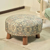 Vintage-inspired round ottoman with intricate patterns, wooden legs, placed on a beige rug in a cozy, modern living space.