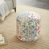 Round floral and paisley patchwork pouf in vibrant multicolor placed on a textured beige rug, with books and a cozy sofa nearby.