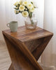 Wooden accent table with an artistic twisted design, holding a glass vase of white flowers and a green ceramic mug, near white curtains.