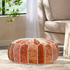 Round patchwork pouf in orange, red, and beige tones with white stitching, placed on a rug near a beige sofa and green plant by a window.