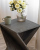 Gray wooden accent table with a twisted design, adorned with a glass vase of white flowers and a green ceramic mug, near white curtains.