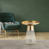 Golden round-top table with a clear glass base, set in a modern living room with a cozy green armchair in the background.