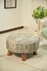 Round ottoman with a vintage-inspired floral pattern, wooden legs, and a cozy setting featuring a beige armchair and green plant.