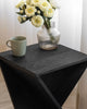 Black wooden accent table with a twisted design, topped with a glass vase of white flowers and a green ceramic mug, near white curtains.