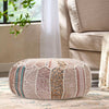 Elegant round pouf in soft pink and beige tones with intricate patchwork design, placed in a cozy living room beside a beige sofa.