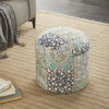 Floral patchwork round pouf in shades of blue and green, resting on a textured beige rug with books and cozy furniture nearby.