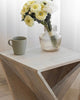 Wooden end table with geometric X-frame, topped with a ribbed glass vase holding yellow and white flowers, next to a green ceramic mug.