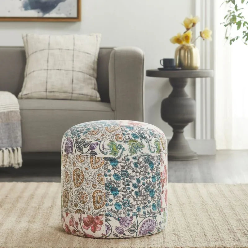 Round pouf with vibrant floral and paisley patchwork in multicolor, placed in a cozy living room with a gray sofa and modern decor.