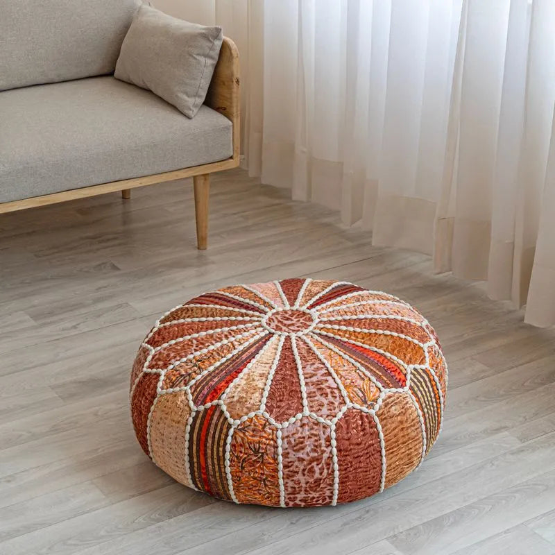 Round patchwork pouf in orange, red, and beige tones with white stitching, placed on a light wood floor near a beige sofa and curtains.