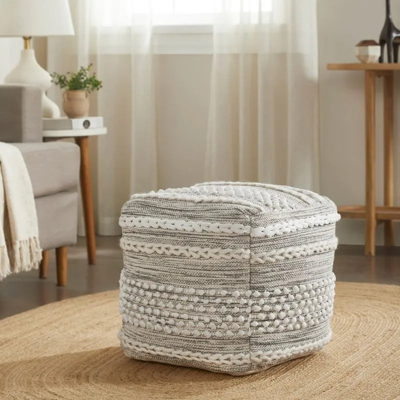 Gray and white textured square pouf with braided and pom-pom patterns, placed on a woven circular rug in a cozy, bright room.