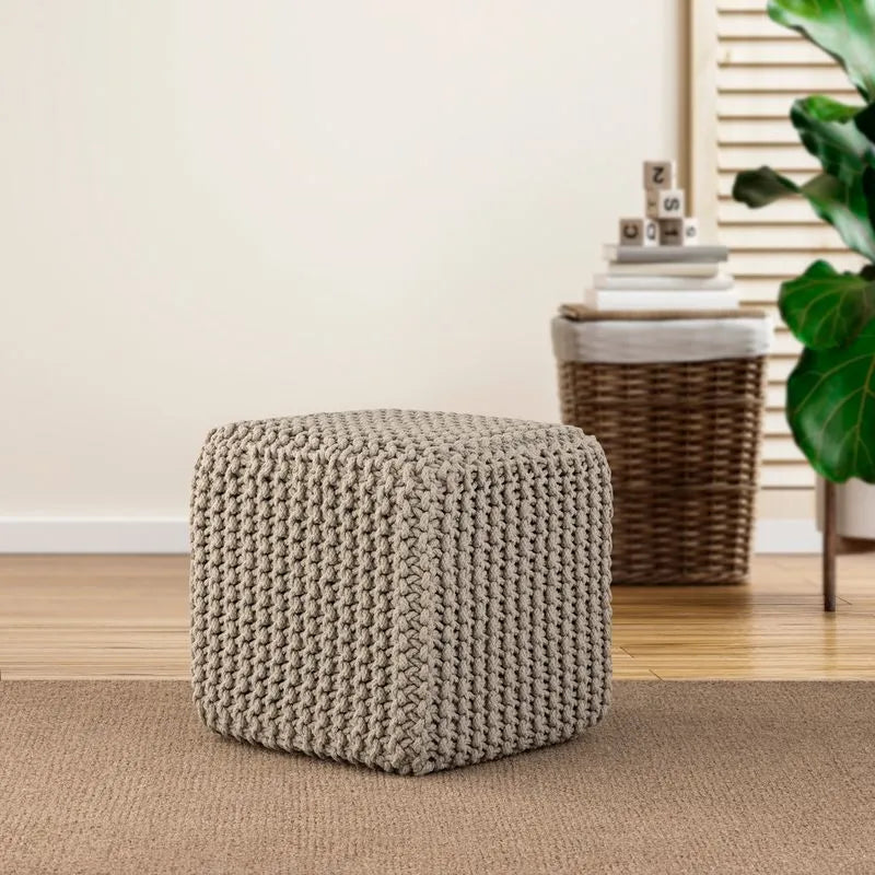 Square beige knitted pouf placed in a cozy living room setting, featuring woven decor and green plants in the background.