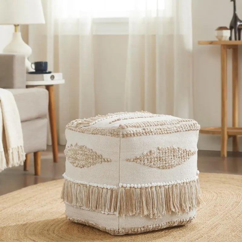 Neutral-toned pouf with textured patterns and fringe detail, placed on a round woven rug in a cozy room with soft natural lighting.