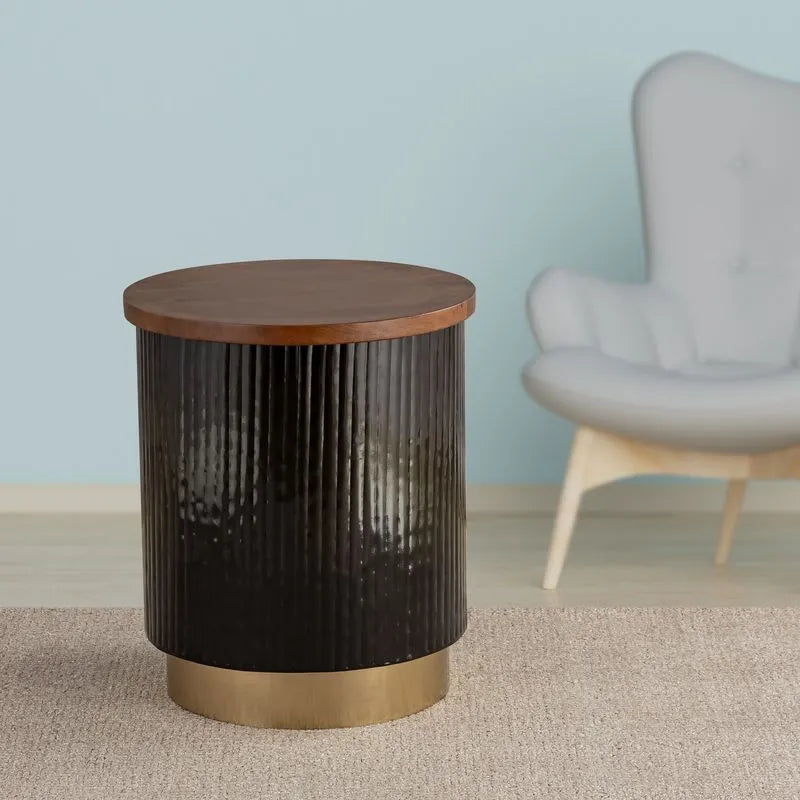 Stylish black cylindrical side table with a textured body, wooden top, and gold metal base, placed beside a modern armchair.